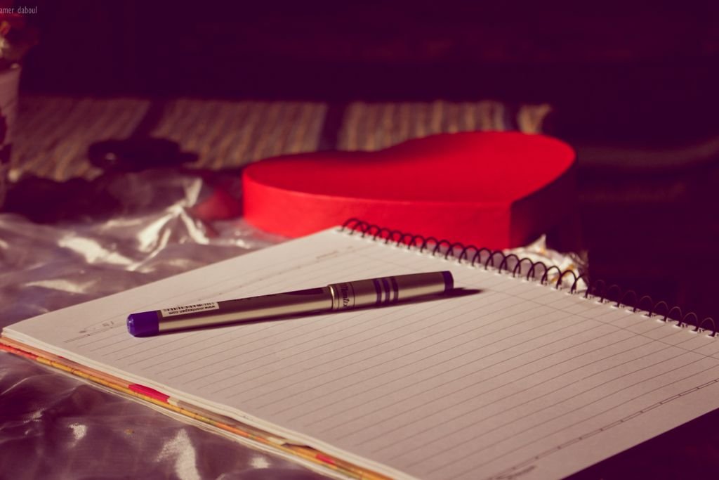 A spiral notebook with a pen placed on a heart-shaped box, symbolizing love and writing.